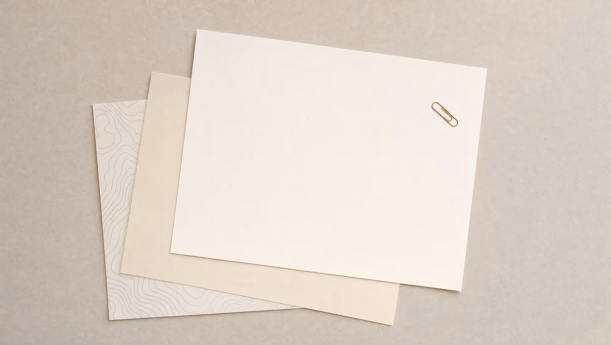 Overhead of overlapping blank warm-paper sheets with faint contour linework and a single gold paperclip on a neutral desk.